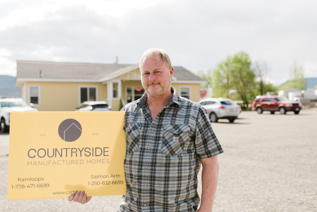 frank ambler co owner of countryside manufactured homes in kamloops stands in front of a showhome holding the countryside sign